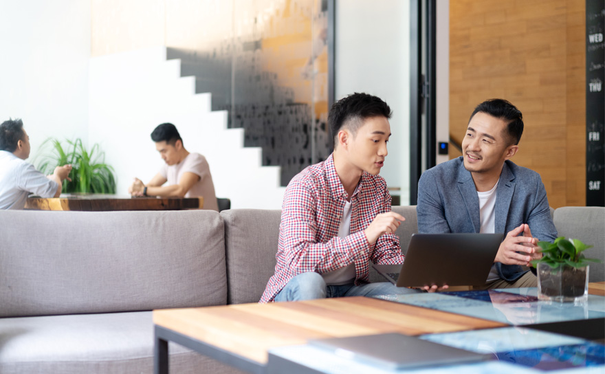 Two men talk on a sofa while looking at a laptop